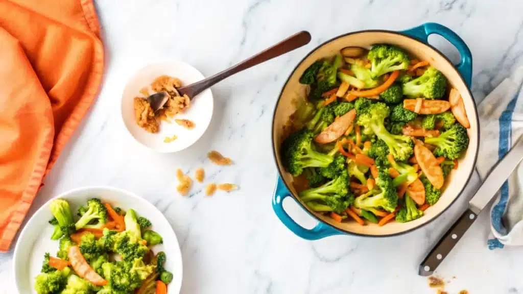 "A colorful dish of sausage broccoli stir-fry in a blue pot, served alongside a bowl of stir-fry and a small dish with a spoon containing a seasoning paste. The meal is garnished with sliced carrots, creating a vibrant presentation