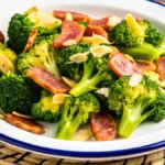 A plate of vibrant sausage broccoli stir-fry, featuring golden-brown sausage slices and fresh green broccoli florets garnished with almond slices, served on a white dish with a blue rim.