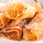 A close-up of golden-brown peach cobbler egg rolls dusted with powdered sugar on a white plate.