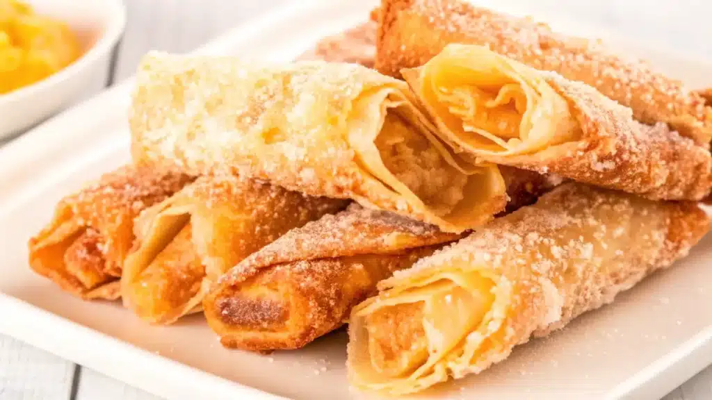 A close-up of golden-brown peach cobbler egg rolls dusted with powdered sugar on a white plate.